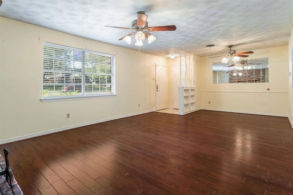 Living area featuring wood-finish flooring, two ceiling fans with integrated lighting, a mirrored wall accent, a built-in shelving partition, and a large multi-pane window