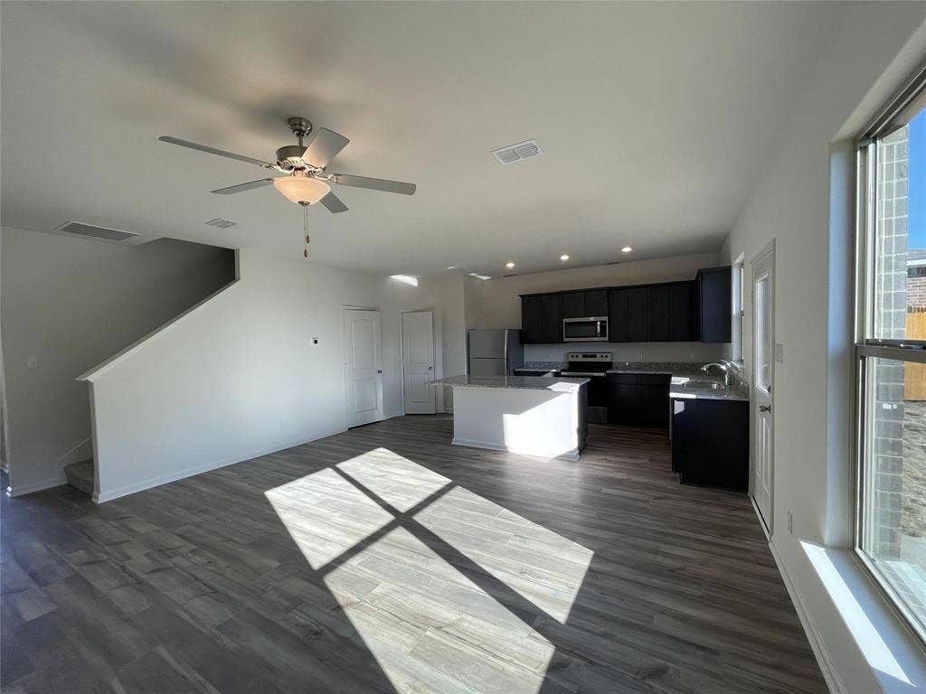 Open-concept floor plan featuring wood-finish flooring, a kitchen with dark cabinetry, a central island with stone-finish countertops, recessed lighting, and a ceiling fan