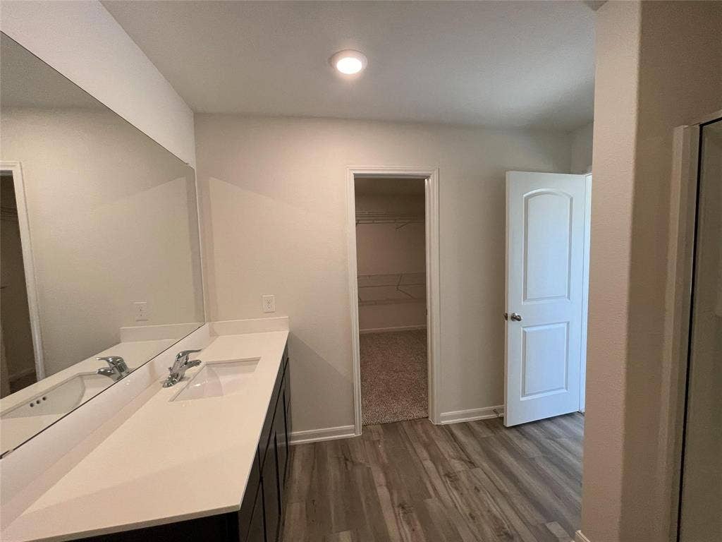 Bathroom featuring a dual-sink vanity with a solid-surface countertop, full-width wall mirror, wood-finish flooring, and a walk-in closet