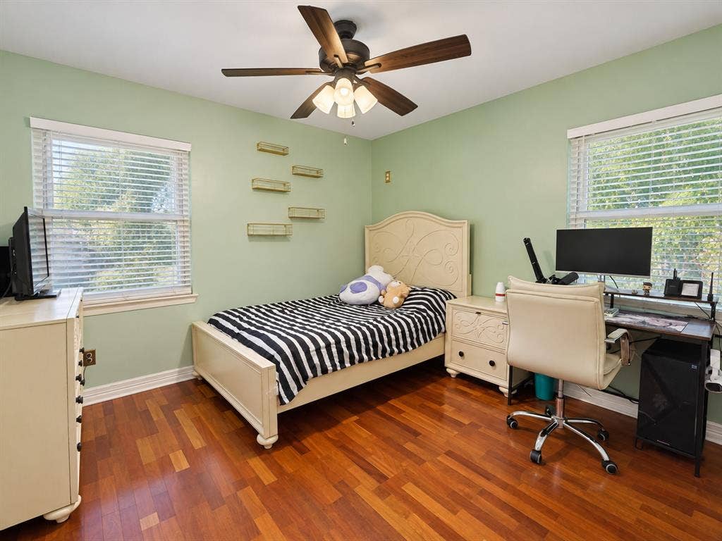 Room featuring wood-finish flooring, two windows with horizontal blinds, built-in wall shelving, and a ceiling fan with integrated lighting