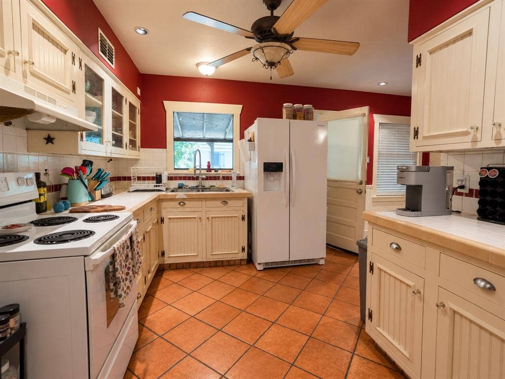 Kitchen featuring cream-colored beadboard cabinetry, terra-cotta tile flooring, tile countertops, glass-front upper cabinet doors, and a ceiling-mounted light fixture with a fan