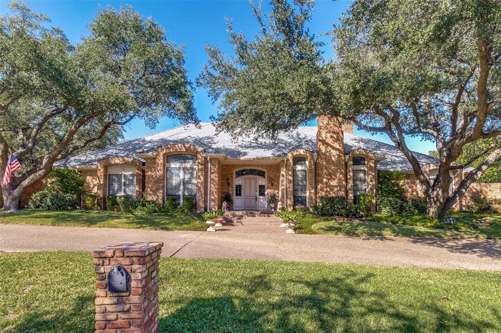 6215 Raintree Court, Dallas, TX 75254 Single-story residence featuring a tan brick exterior, arched window detailing, a chimney, and a concrete driveway bordered by established trees