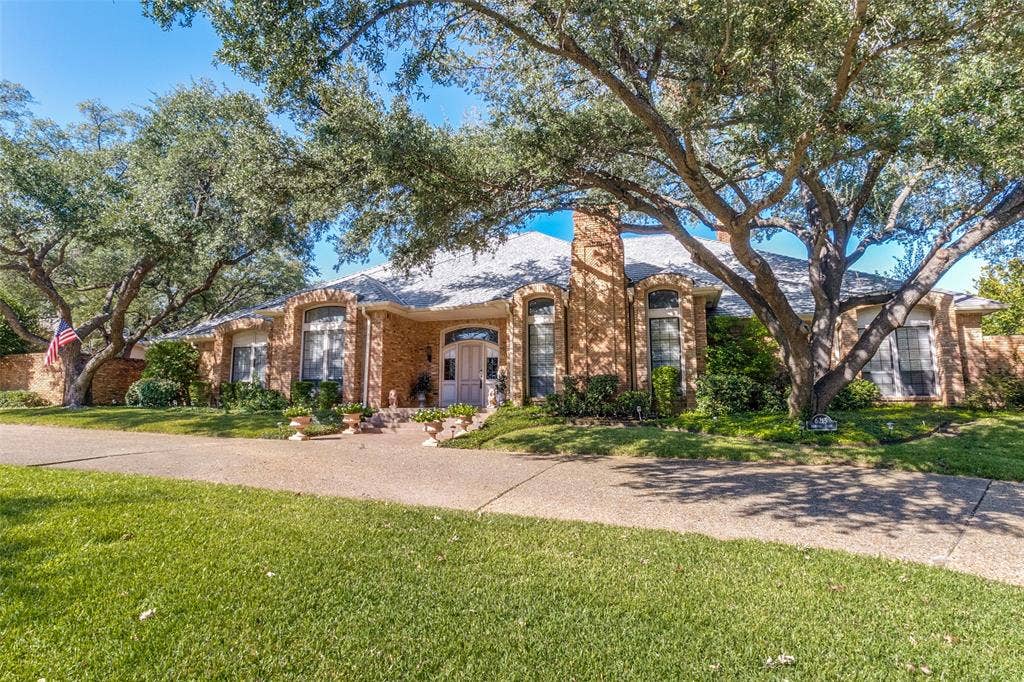 6215 Raintree Court, Dallas, TX 75254 Single-story brick exterior featuring a prominent chimney, arched window casings, mature canopy trees, and a concrete driveway