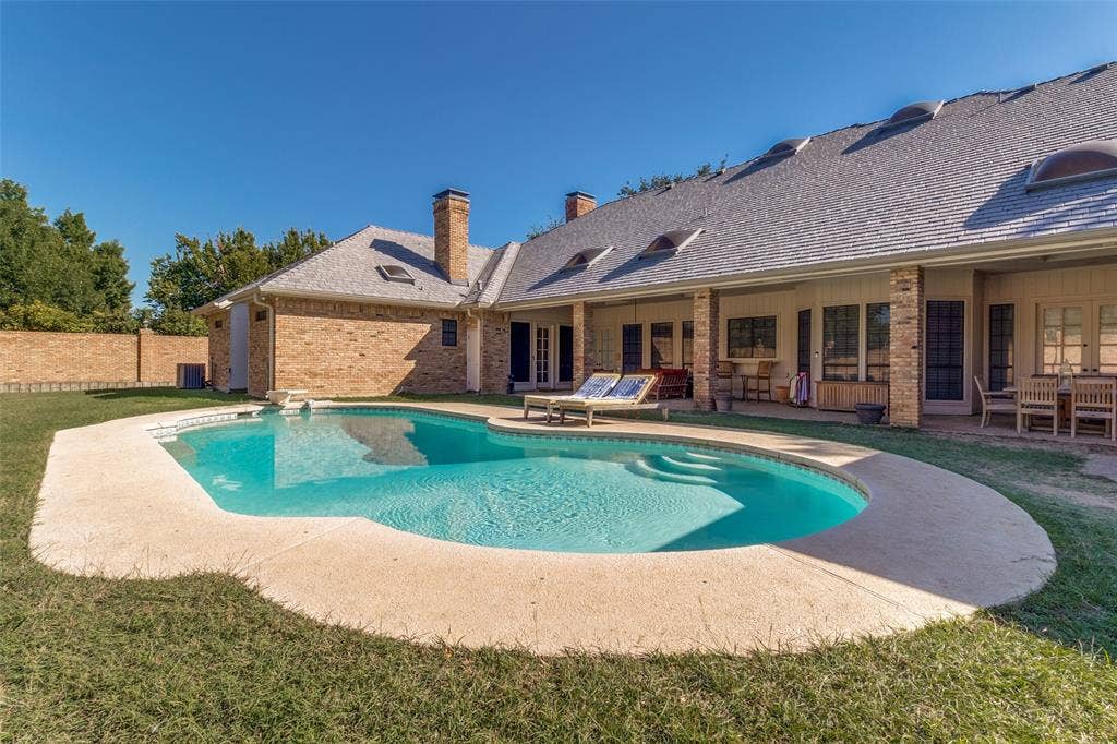 6215 Raintree Court, Dallas, TX 75254 In-ground pool with stone-texture decking, covered patio area, brick exterior, multiple skylights, and a high-pitched roofline