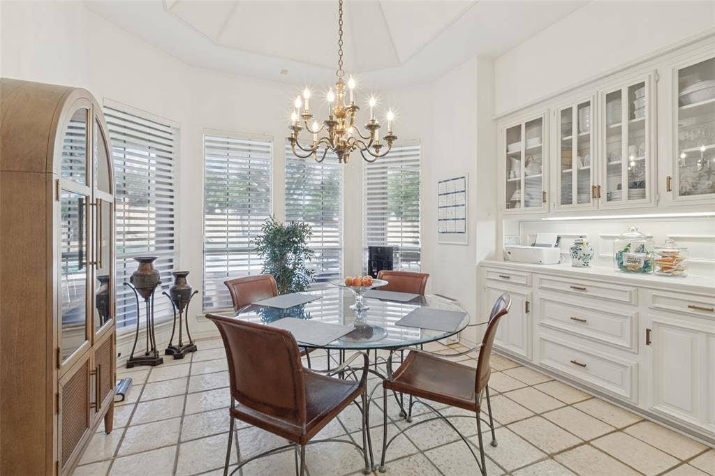 6215 Raintree Court, Dallas, TX 75254 Vaulted ceiling with a multi-arm chandelier, large windows with horizontal blinds, built-in white cabinetry with glass-front display doors, under-cabinet lighting, and light-toned tile flooring