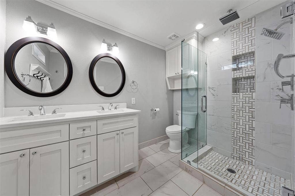 200 Oriental Road, Waco, TX 76710 Dual-sink vanity with white cabinetry and countertop, glass-enclosed walk-in shower featuring decorative tile inlay and built-in shelving, and dual circular wall-mounted mirrors