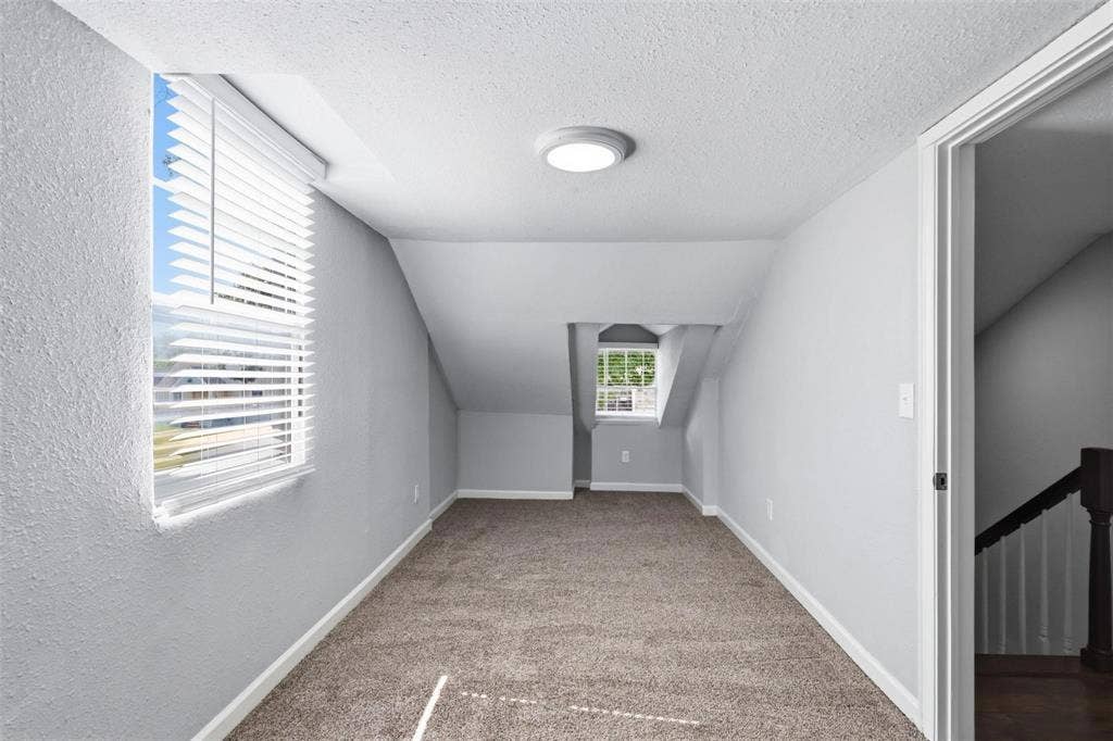 1227 Wilbur Street, Dallas, TX 75224 Neutral-toned interior room featuring slanted ceiling lines, wall-to-wall carpeting, and multiple windows with horizontal blinds