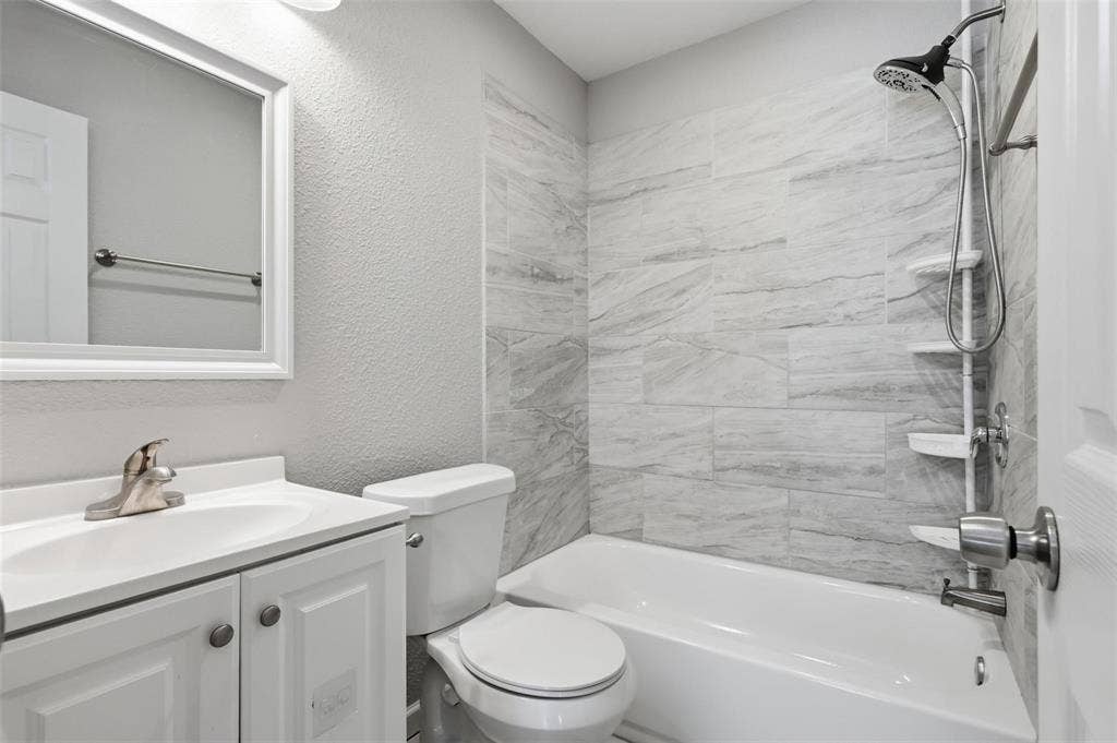 1227 Wilbur Street, Dallas, TX 75224 Bathroom featuring a white vanity cabinet, rectangular undermount sink with a polished faucet, wall-mounted mirror, toilet, and a bathtub with stone-patterned tile surround