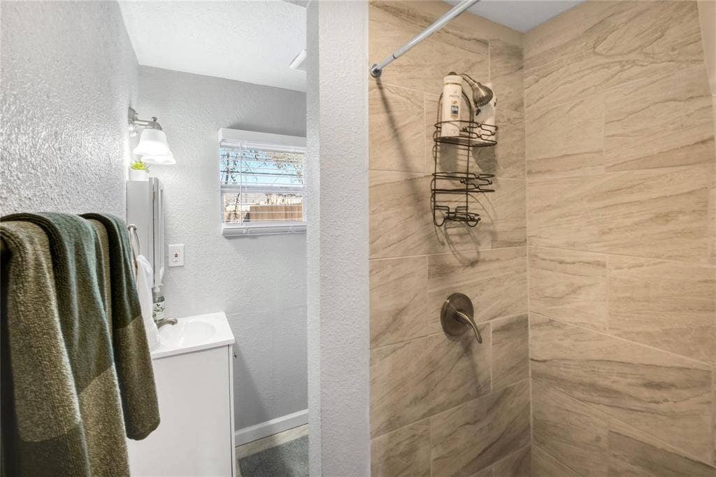 1227 Wilbur Street, Dallas, TX 75224 Bathroom featuring a shower stall with neutral-toned, large-format tile surround, single-handle brushed metal faucet, wall-mounted light fixture, and a white cabinet vanity with a built-in sink