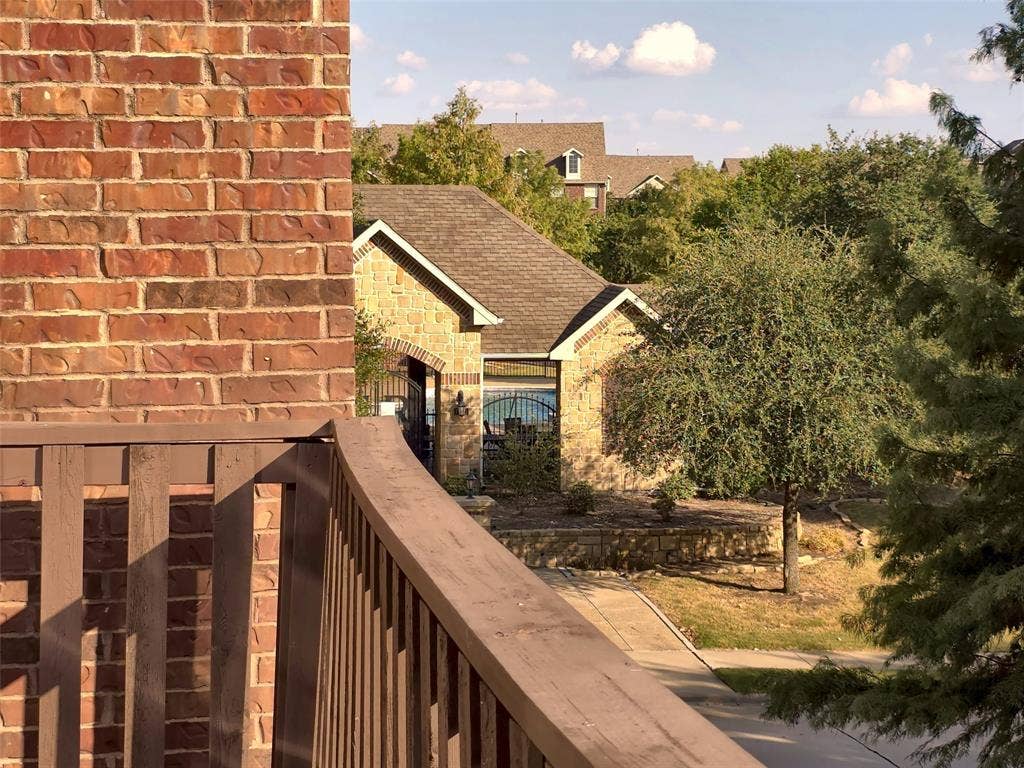 1504 Fox Run Drive, Irving, TX 75063 Exterior balcony featuring a wood-style railing and red brick siding, overlooking a neighboring stone-clad building with a shingled roof and mature landscaping