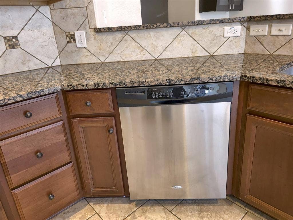 1504 Fox Run Drive, Irving, TX 75063 Stained wood cabinetry, stone-finish countertops, stainless steel dishwasher, and diagonal-pattern tile backsplash