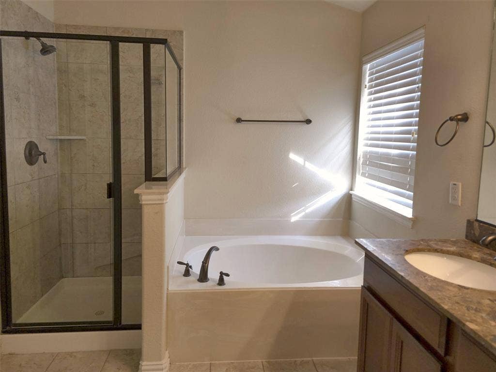 1504 Fox Run Drive, Irving, TX 75063 Walk-in shower with stone-finish tile surround and dark metal framing, soaking tub with deck-mounted faucet, wood-finish cabinetry, stone-composite countertop, and under-mount sink