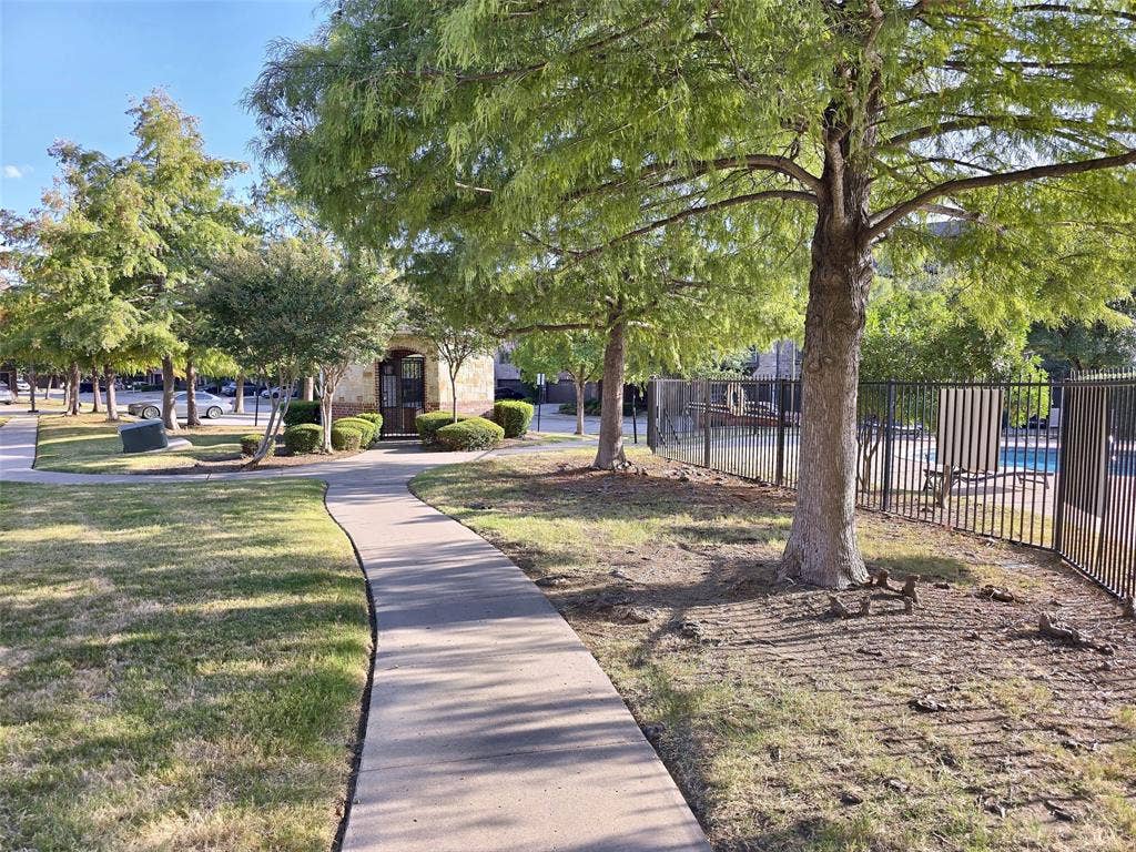 1504 Fox Run Drive, Irving, TX 75063 Concrete walking path bordered by mature trees, manicured lawn, established landscaping, and black metal fencing surrounding a pool area