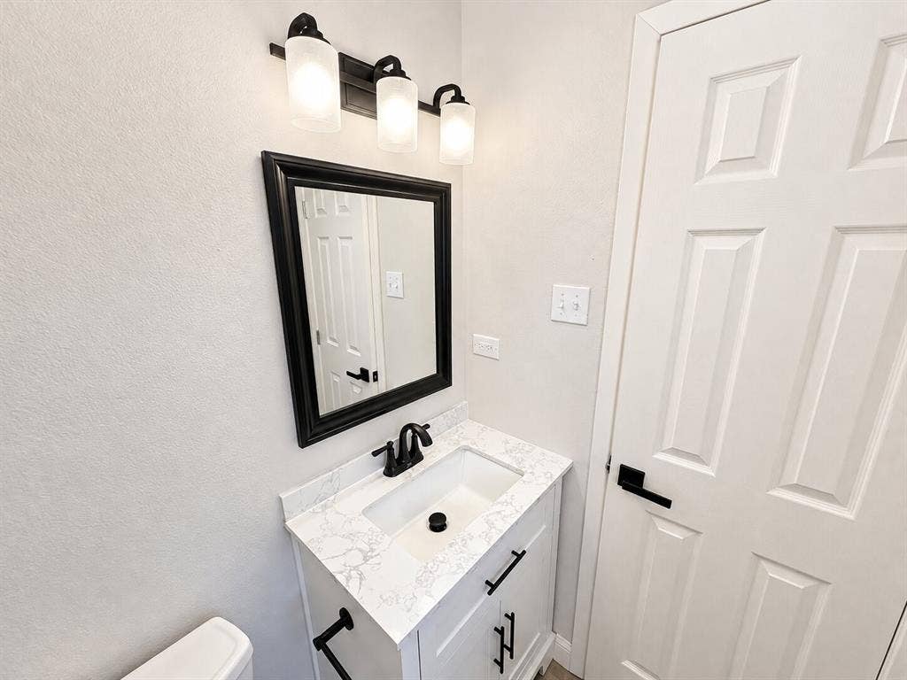 1307 Crestridge Drive, Plano, TX 75075 Vanity featuring a white cabinet, stone-finish countertop, and integrated rectangular sink with matte black hardware, accompanied by a framed mirror, three-light fixture with frosted glass shades, and a six-panel door