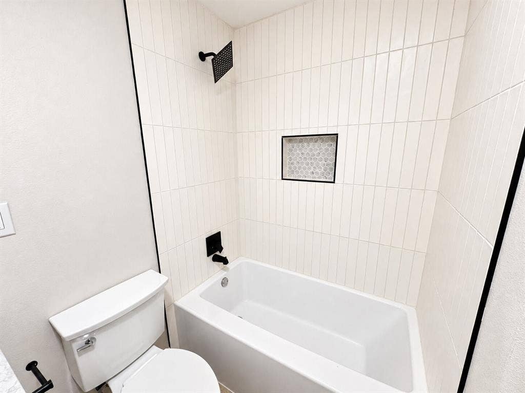 1307 Crestridge Drive, Plano, TX 75075 Bathtub alcove featuring vertical white wall tile, matte black shower fixtures, a recessed niche with decorative tile inlay, and a white toilet