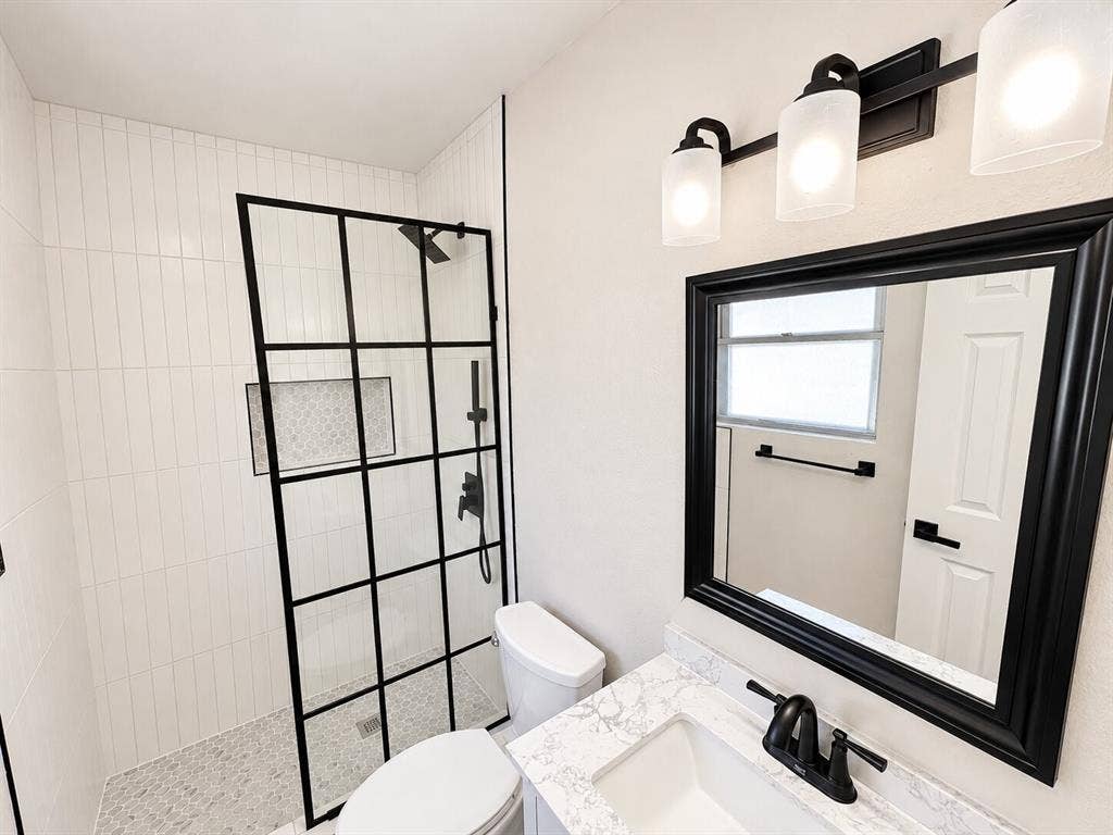 1307 Crestridge Drive, Plano, TX 75075 Bathroom featuring a glass shower enclosure with black grid framing, vertical wall tile, stone-patterned countertop, vanity mirror with black frame, and a three-light wall-mounted fixture