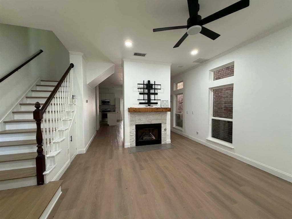5864 Oram Street, Dallas, TX 75206 Interior space featuring wood-finish flooring, a central fireplace with stone detailing and a timber mantel, tall windows with brick exterior accents, and a staircase with a dark wood handrail
