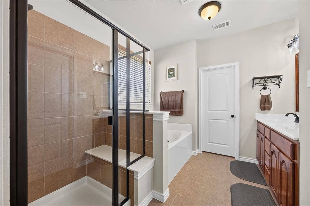 1514 Natalie Lane, Duncanville, TX 75137 Walk-in shower featuring a built-in bench, tiled surround, and a dark-finish frame, paired with a white soaking tub, wood-finish cabinetry, and a countertop with an integrated sink