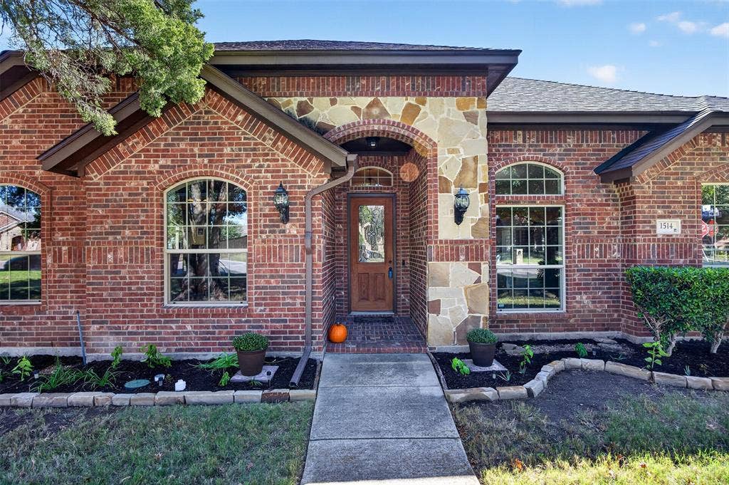1514 Natalie Lane, Duncanville, TX 75137 Brick exterior featuring stone masonry detailing, arched window accents, wood-finish entry door with glass insert, and tiered landscaping beds