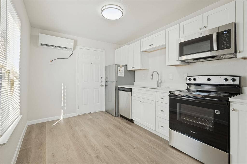 Kitchen featuring white shaker-style cabinetry, wood-finish flooring, stainless steel appliances, and a wall-mounted ductless heating and cooling unit
