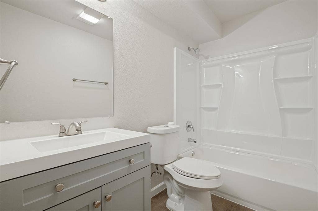 Bathroom featuring a grey shaker-style vanity, white countertop with integrated sink, wall-mounted mirror, and a tub-shower combination with built-in shelving