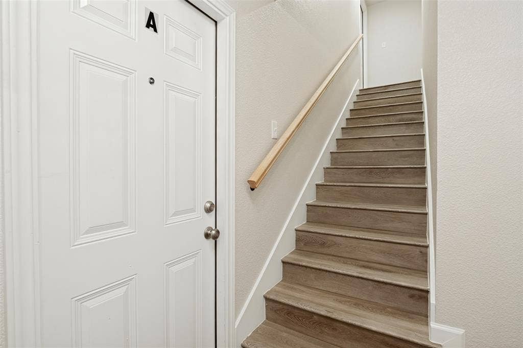 White paneled door with integrated deadbolt and peephole, accompanied by a staircase featuring wood-finish treads and a light-toned wood handrail