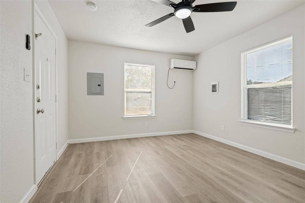 Neutral-toned space featuring wood-finish flooring, a wall-mounted ductless mini-split HVAC unit, two windows with horizontal blinds, and a ceiling fan with integrated lighting