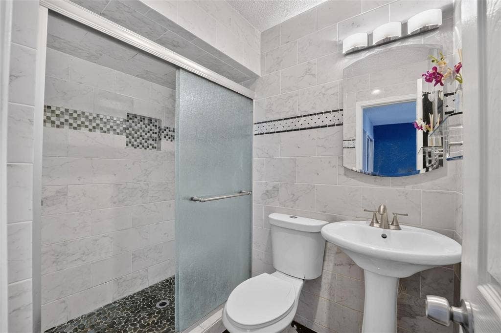 2130 Bowie Drive, Carrollton, TX 75006 Full bathroom featuring a shower with a sliding glass door, stone-finish tile wall surround, pebble-style flooring, a pedestal sink, and a three-bulb wall-mounted light fixture