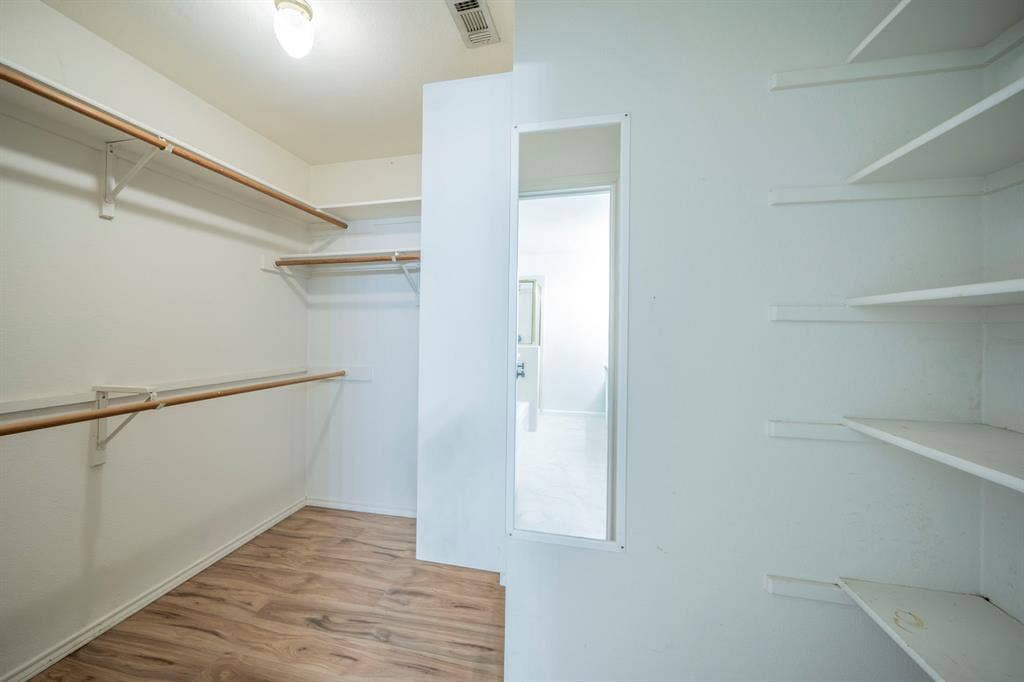Walk-in storage space featuring wood-finish flooring, white built-in shelving, hanging rods, and a full-length wall-mounted mirror