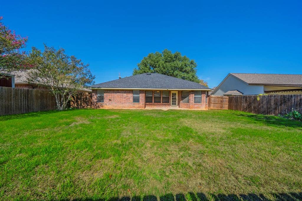 Expansive backyard featuring a large grass lawn, brick exterior, wood privacy fencing, and mature trees