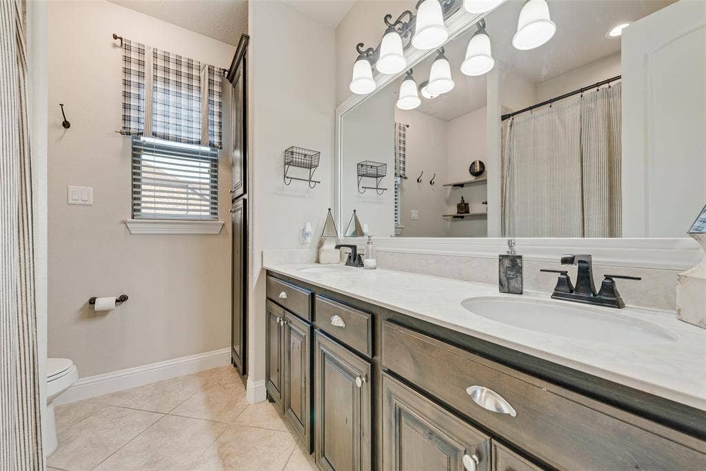7231 Judy Drive, Ovilla, TX 75154 Dual-sink vanity featuring stone-finish countertops, dark-stained cabinetry with recessed-panel doors, matte black fixtures, and a large framed wall mirror