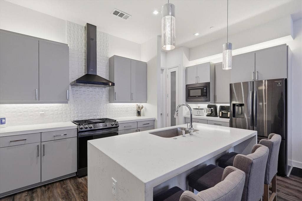 1813 Park Avenue, #106, Dallas, TX 75215 Modern kitchen featuring grey flat-panel cabinetry, quartz countertops, a central island with an integrated sink, textured tile backsplash, and stainless steel appliances