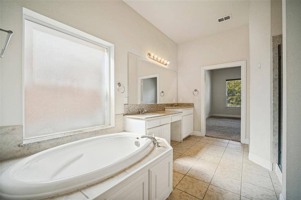 Bathroom featuring a drop-in soaking tub with a tiled surround, dual-sink vanity with stone-style countertops, expansive wall-mounted mirror, and tile flooring