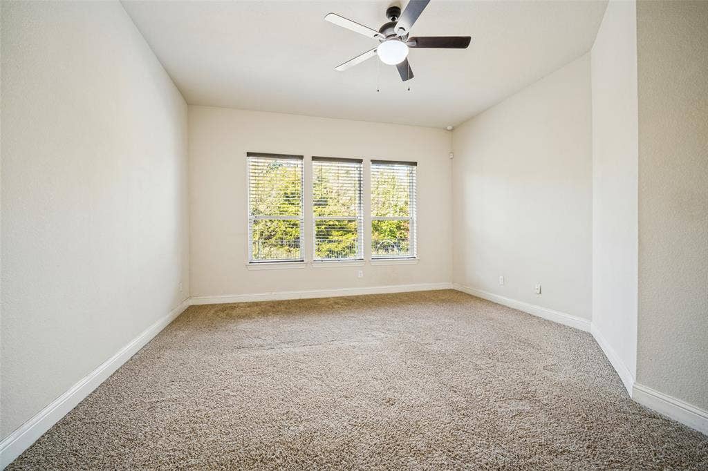 Neutral-toned walls, high-pile carpeting, triple window configuration with integrated blinds, and a central ceiling fan with an integrated light fixture