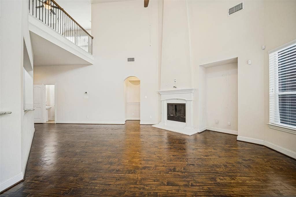 Double-height ceiling, fireplace with detailed mantel, wood-finish flooring, and an elevated mezzanine with a wrought iron railing