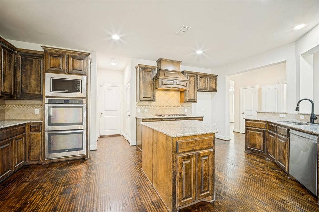Kitchen featuring stained wood cabinetry, granite countertops, integrated double wall ovens, built-in range hood, and wood-finish flooring