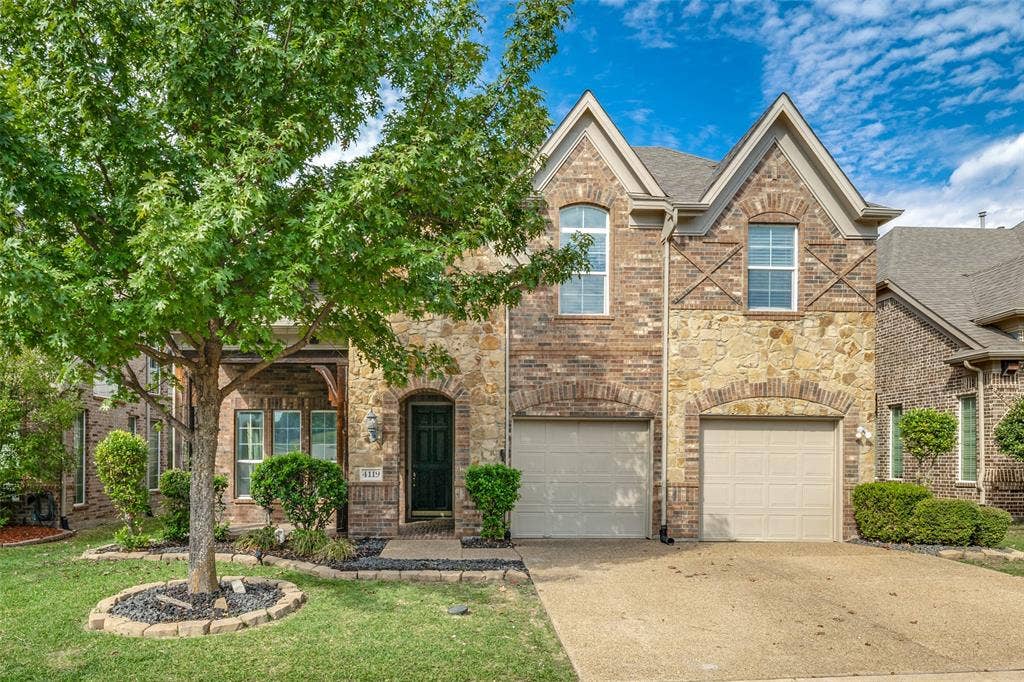 Two-story exterior featuring multi-textured brick and stone masonry, arched window detailing, double gabled roofline, and an attached two-car garage