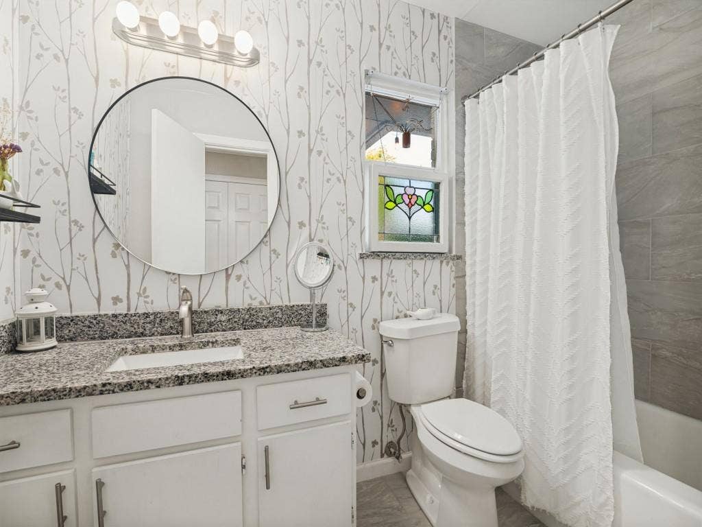 200 S Weatherred Drive, Richardson, TX 75080 Bathroom featuring a vanity with stone-finish countertop, round mirror, patterned wall covering, window with decorative stained-glass inlay, and shower-tub combination with grey tile surround