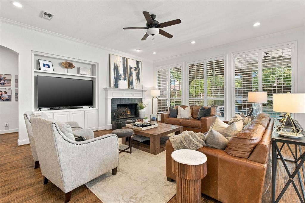 Living area featuring wood-finish flooring, a built-in media cabinet, a stone-surround fireplace with a classic mantel, recessed lighting, and multiple windows with plantation shutters