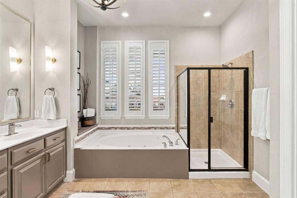 Jetted soaking tub with tile surround, walk-in shower featuring a black metal frame, cabinetry with metallic hardware, and plantation shutters