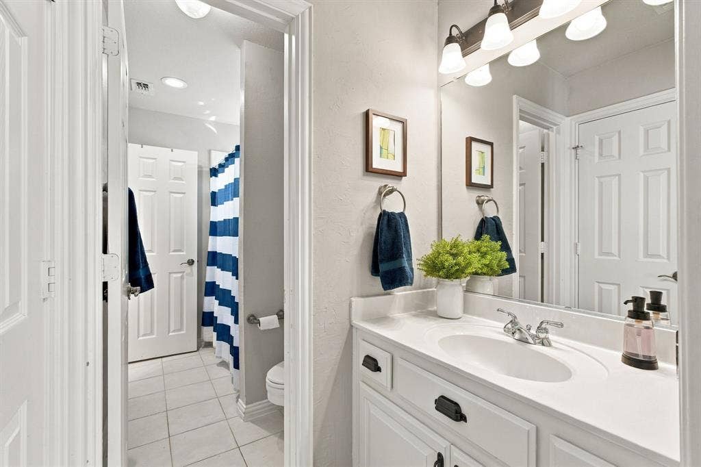 Bathroom featuring a white vanity with matte black hardware, integrated sink with chrome faucet, large wall-mounted mirror, multi-bulb overhead vanity lighting, and light-toned tile flooring