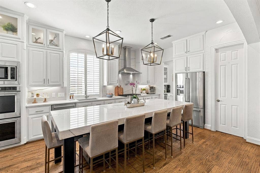 Kitchen featuring white cabinetry with glass-front uppers, an expansive central island with stone-surface countertop, stainless steel appliances, geometric pendant lighting, and wood-finish flooring