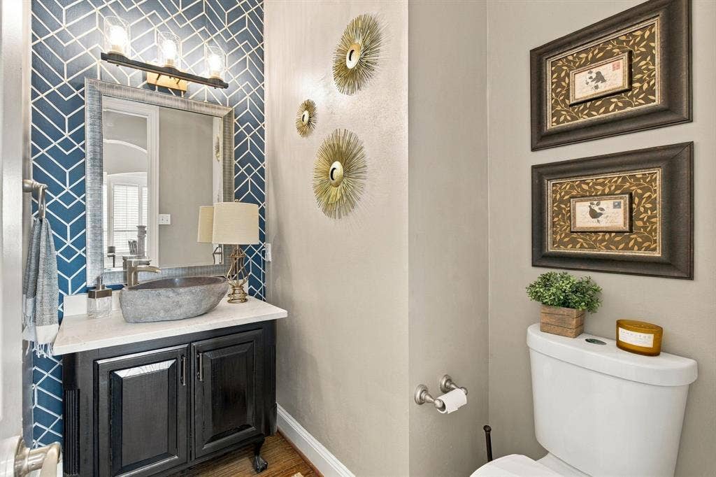 Black vanity cabinet with a light-toned countertop, stone-finish vessel sink, geometric patterned accent wall, and three-light vanity fixture