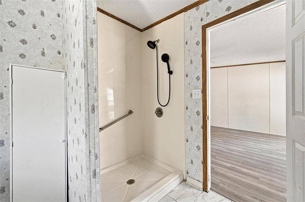 Walk-in shower enclosure featuring smooth-surface surround panels, a handheld showerhead, a metal grab bar, and wood-finish flooring in the room beyond