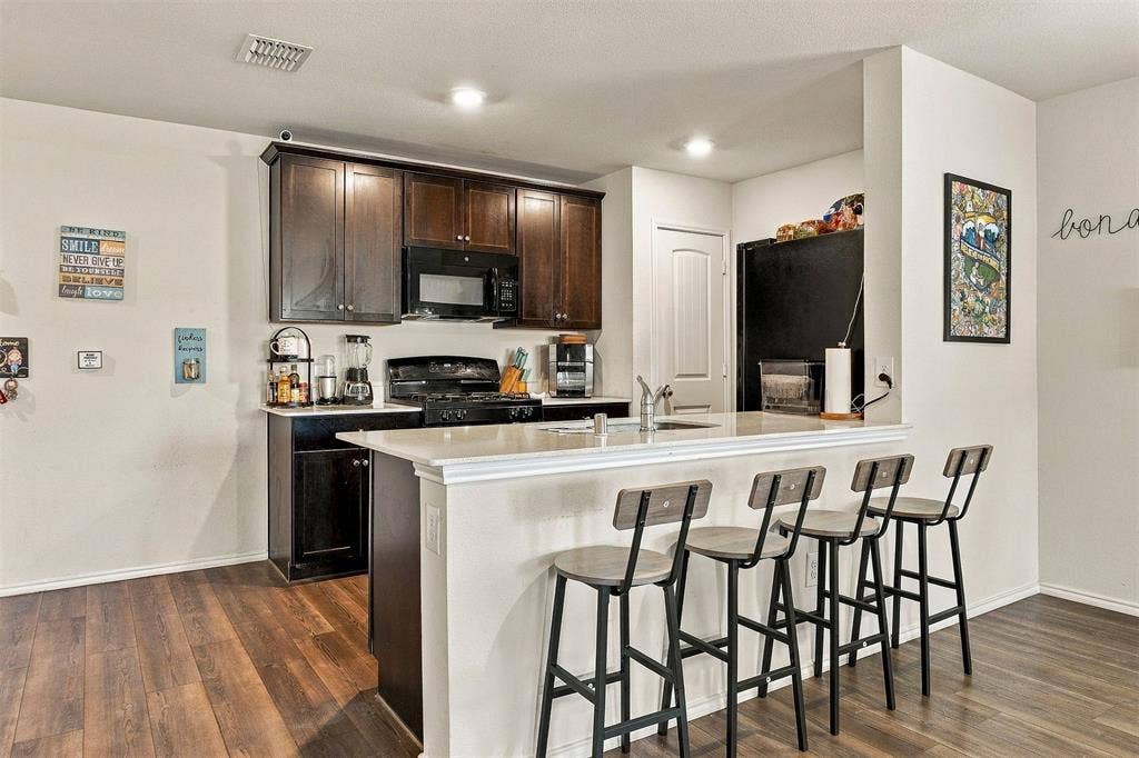 1516 Stonefield Drive, Anna, TX 75409 Kitchen featuring dark wood cabinetry, a light-toned stone-surface peninsula, wood-finish flooring, and built-in black appliances