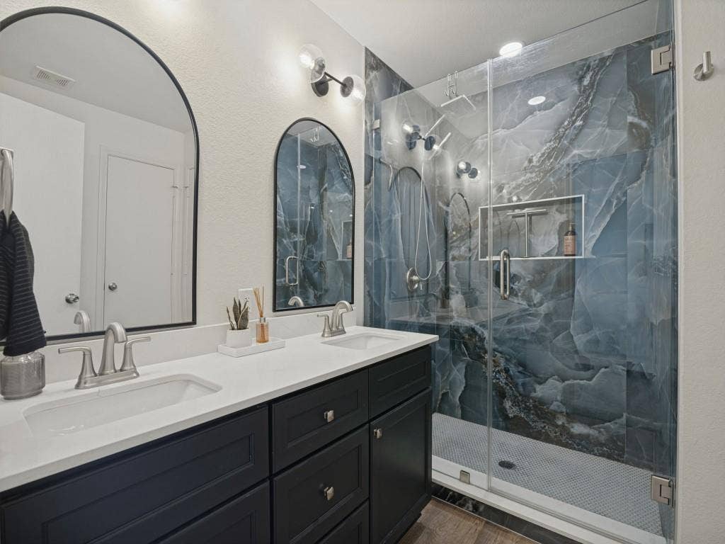 1728 Harvest Crossing Drive, Wylie, TX 75098 Dual vanity with solid-surface countertop and dark cabinetry, arched wall mirrors, and walk-in glass-enclosed shower featuring large-format patterned wall tiles and a built-in recessed niche