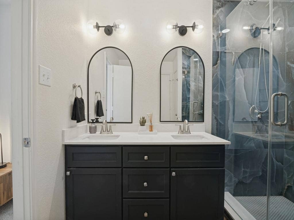 1728 Harvest Crossing Drive, Wylie, TX 75098 Double vanity featuring charcoal-colored cabinetry, dual undermount sinks, white solid-surface countertop, dual arched wall mirrors, and modern two-light fixtures