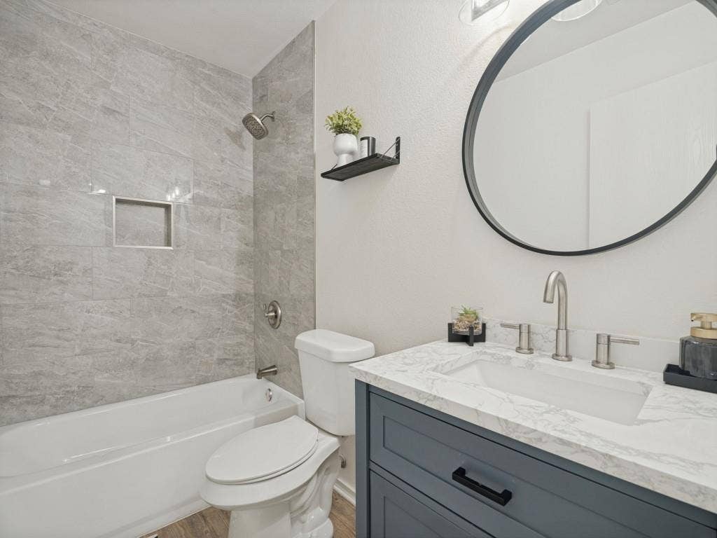 1728 Harvest Crossing Drive, Wylie, TX 75098 Bathroom featuring a vanity with shaker-style cabinetry, stone-finish countertop, integrated rectangular sink, wall-mounted circular mirror, and tub-shower combination with grey tile surround and built-in recessed niche