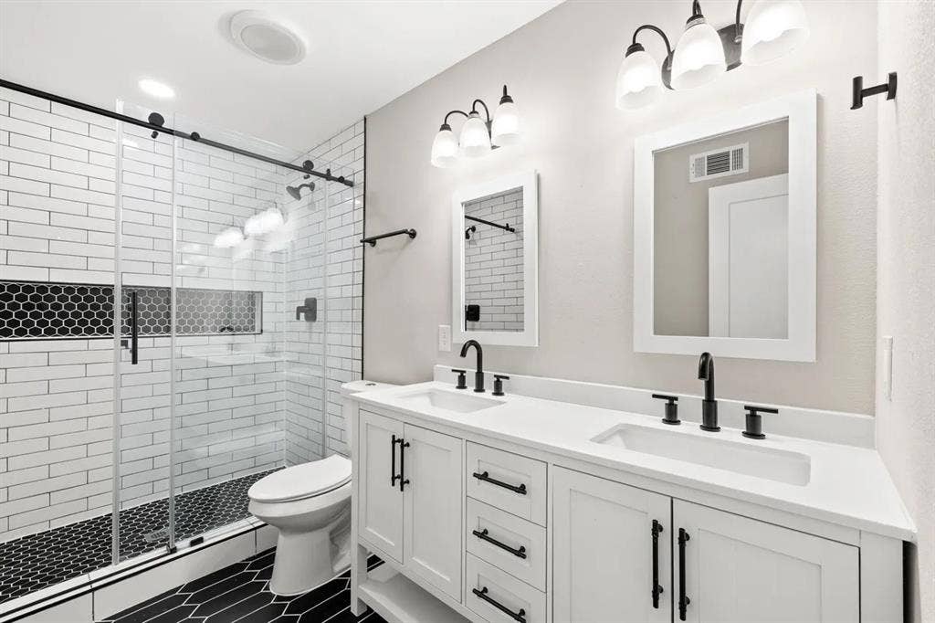 Dual-sink vanity with a white solid-surface countertop and matte black hardware, walk-in shower featuring a glass enclosure with sliding track, white subway wall tile, and hexagonal black floor tile