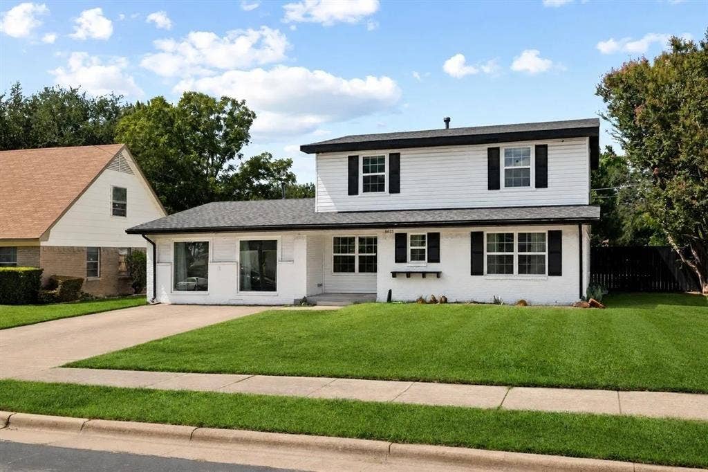 Two-story residence featuring white painted brick and siding, a dark shingled roof, contrast shutters, and an expansive front lawn